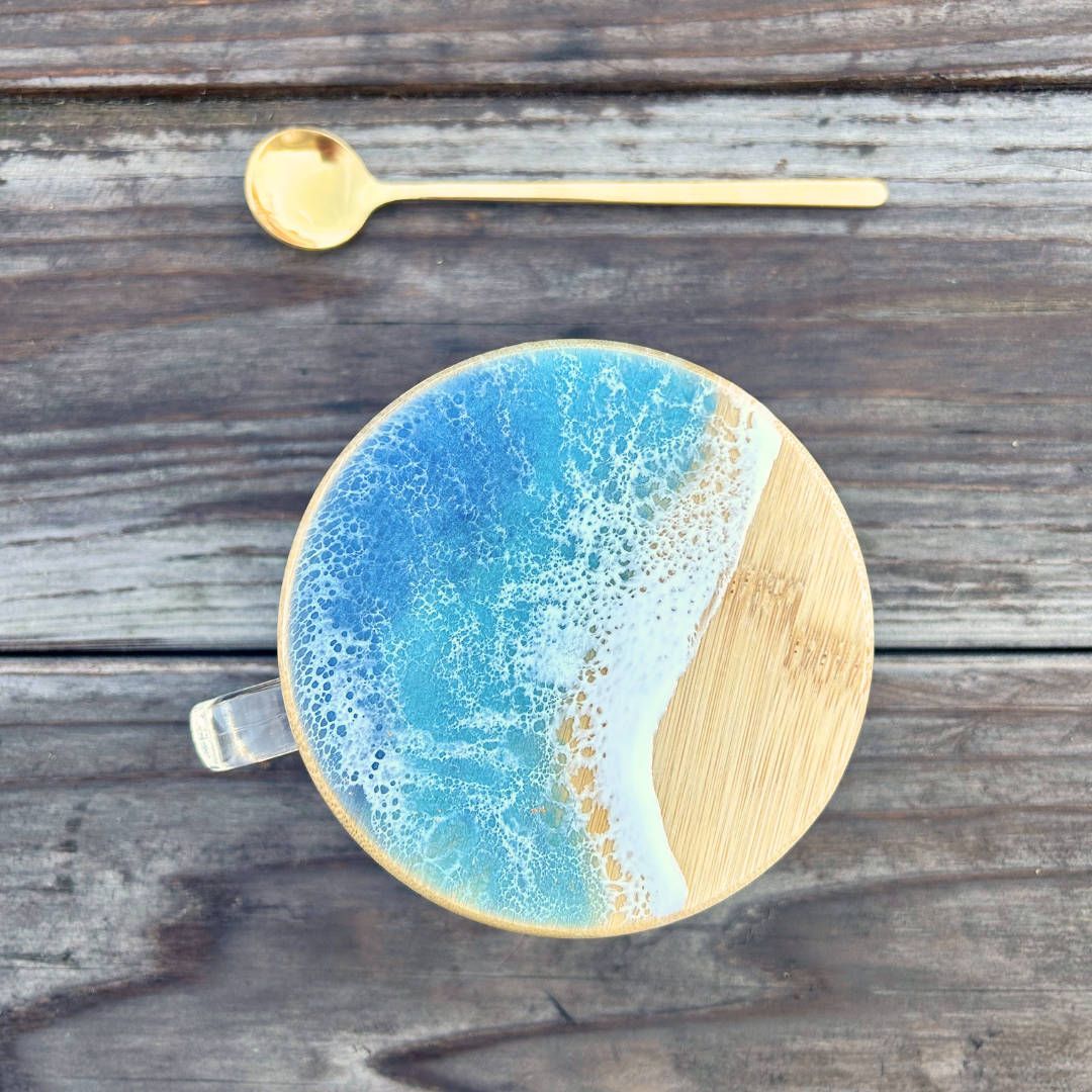 A top-down view of a round bamboo cup with a blue and white foam-patterned lid, placed on a rustic gray wooden table with a gold spoon above it.